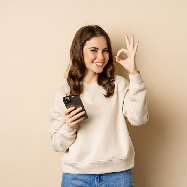 Beautiful woman holding cellphone, mobile phone and okay sign, recommending application, shopping app, standing over beige background.